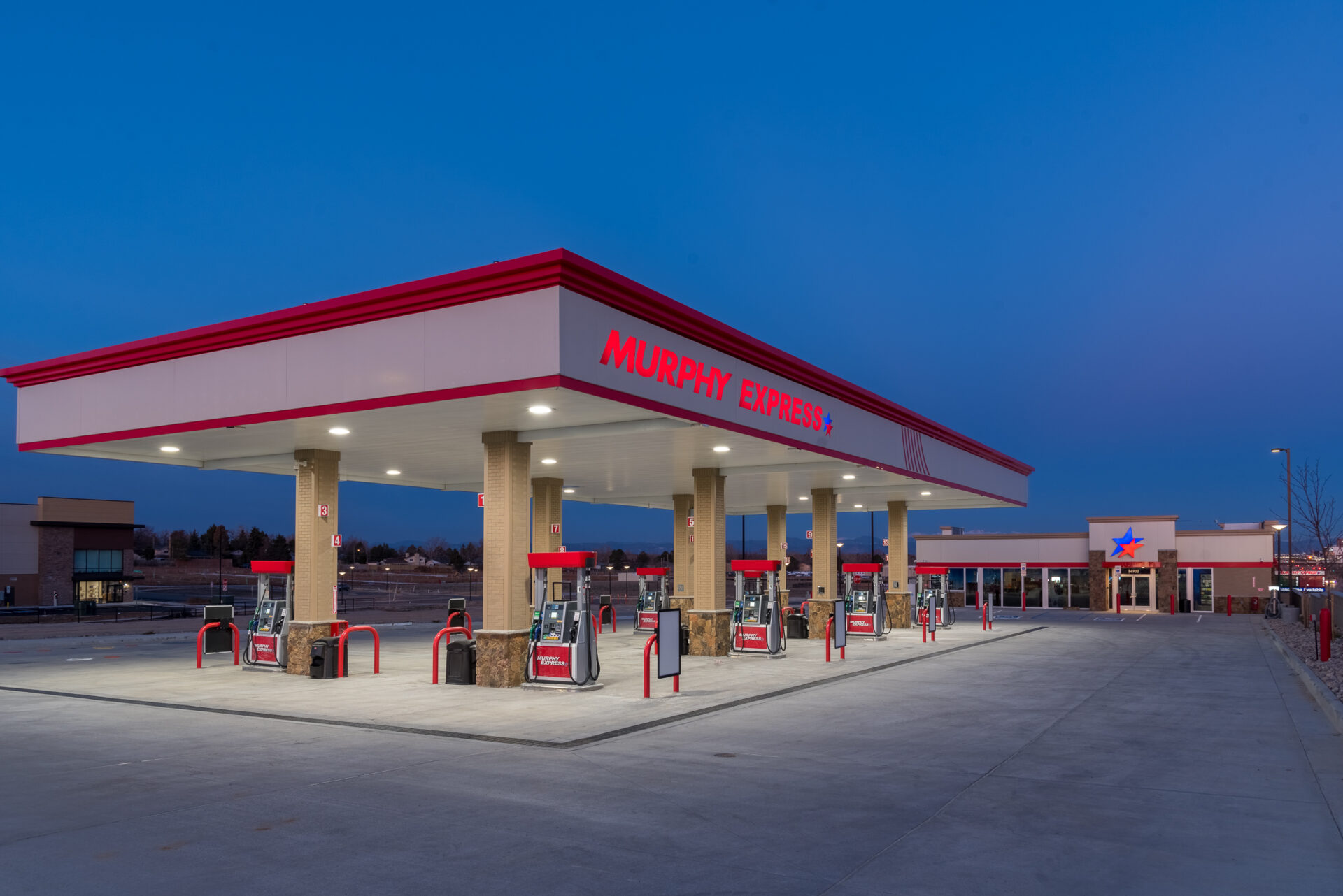 A well-lit Murphy Express gas station at dusk, featuring multiple fuel pumps under a canopy with red accents, with a convenience store in the background. The sky is a deep blue, hinting at twilight, and the ground appears clean and spacious.