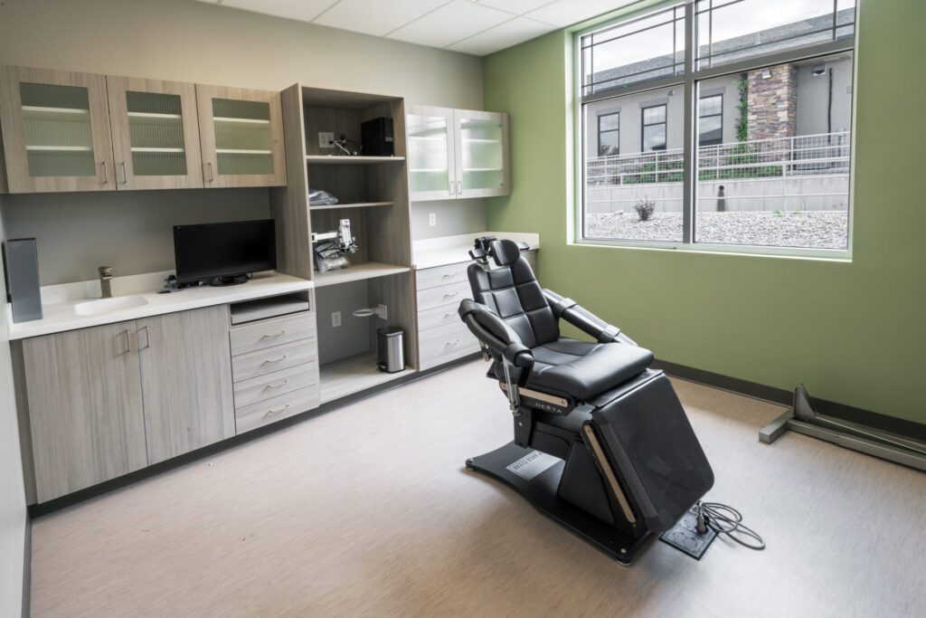 A modern medical examination room with light wood cabinets and shelves, a black reclining exam chair in the center, and a computer on the counter. The room has green walls, a large window offering natural light, and a clean, organized appearance.