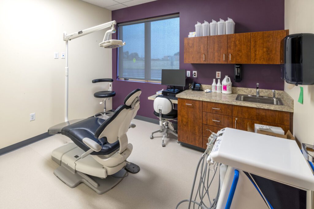 A modern dental exam room with a dentist chair, overhead light, and equipment tray on the left. On the right, there is a counter with a sink, soap dispenser, computer, and various supplies. The room has beige walls, a large window, and purple accent walls.