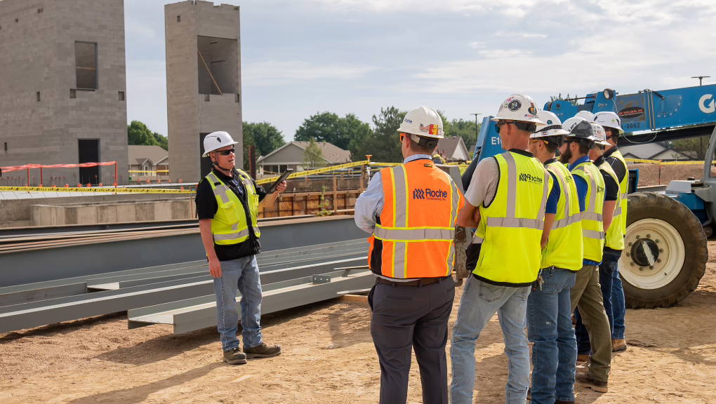 A construction supervisor in a yellow vest and hard hat addresses workers in safety gear at a commercial construction site, highlighting the importance of teamwork with your dedicated commercial construction partner amid steel beams and unfinished structures.