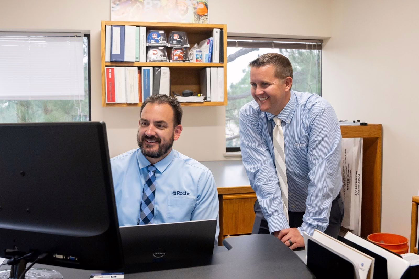 Two men in dress shirts and ties smile while working together at a computer, collaborating on preconstruction planning that saves time. One is seated at the desk using the computer, while the other stands beside him, leaning in to look at the screen.