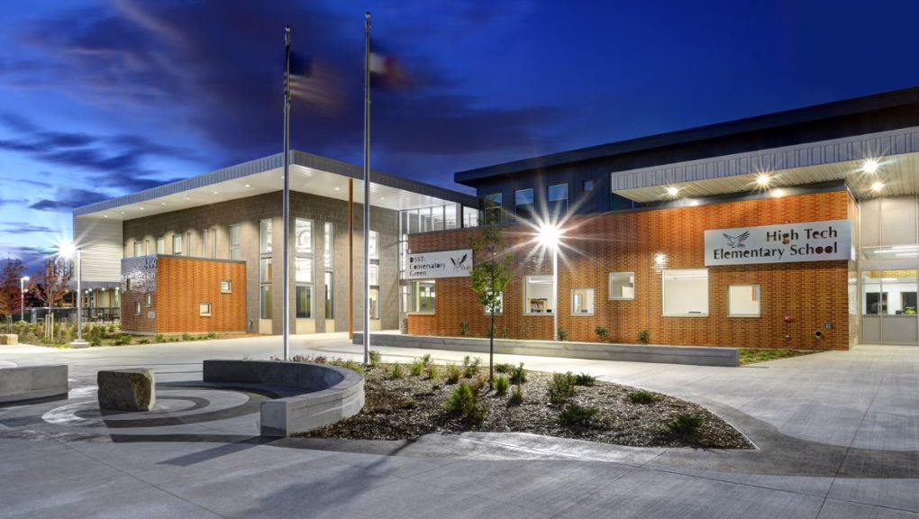 Modern school building at dusk with illuminated windows, a circular concrete bench, landscaping, and two flagpoles in front. Signs on the building read "High Tech Elementary School.