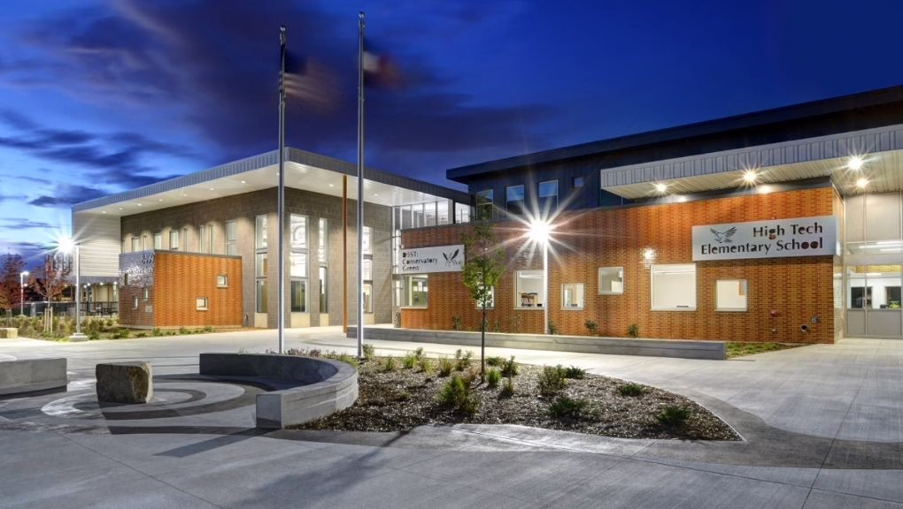 A modern brick and glass school building at dusk with lit windows, two flagpoles, landscaped grounds, round bench seating, and signs reading "High Tech Elementary School" near the main entrance.