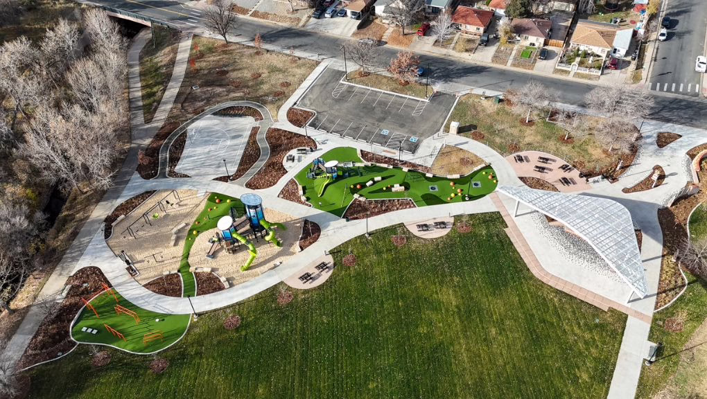 Aerial view of a city park with a playground featuring slides, climbing structures, shaded seating areas, pathways, a parking lot, grass fields, trees, and nearby houses.